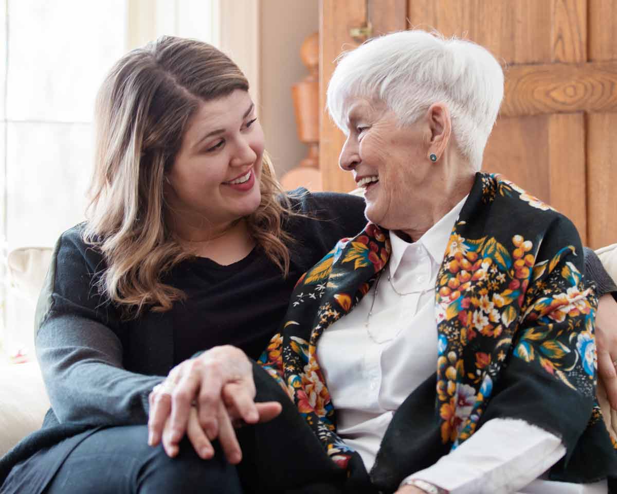 Young woman and older woman sit closely together and smile at each other