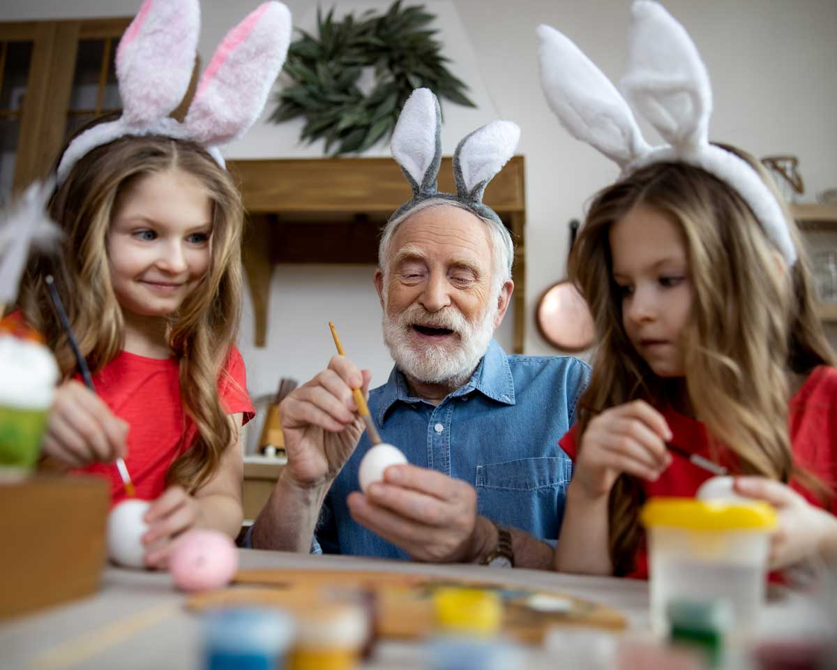 Older man and two young girls wear bunny ears and dye Easter eggs together at table