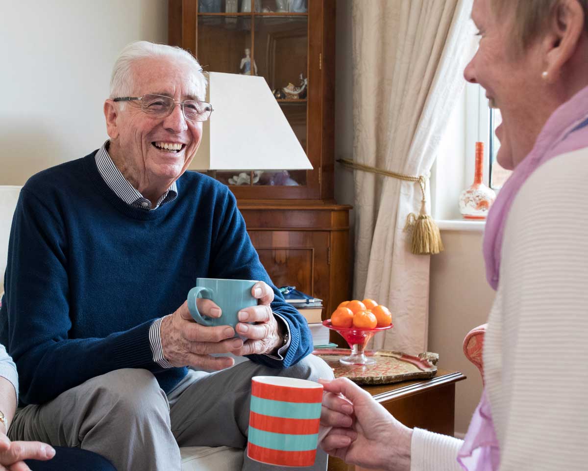 Older male and female sit on couches while smiling and holding coffee mugs