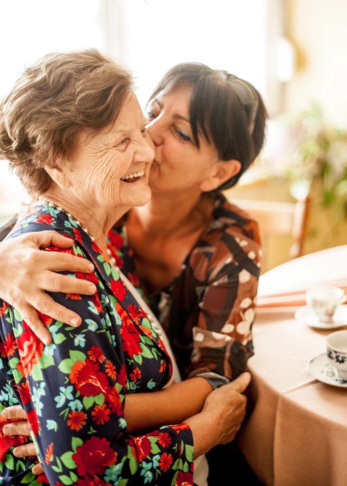 Older woman sits at dining table and smiles while receving kiss on cheek from female family member