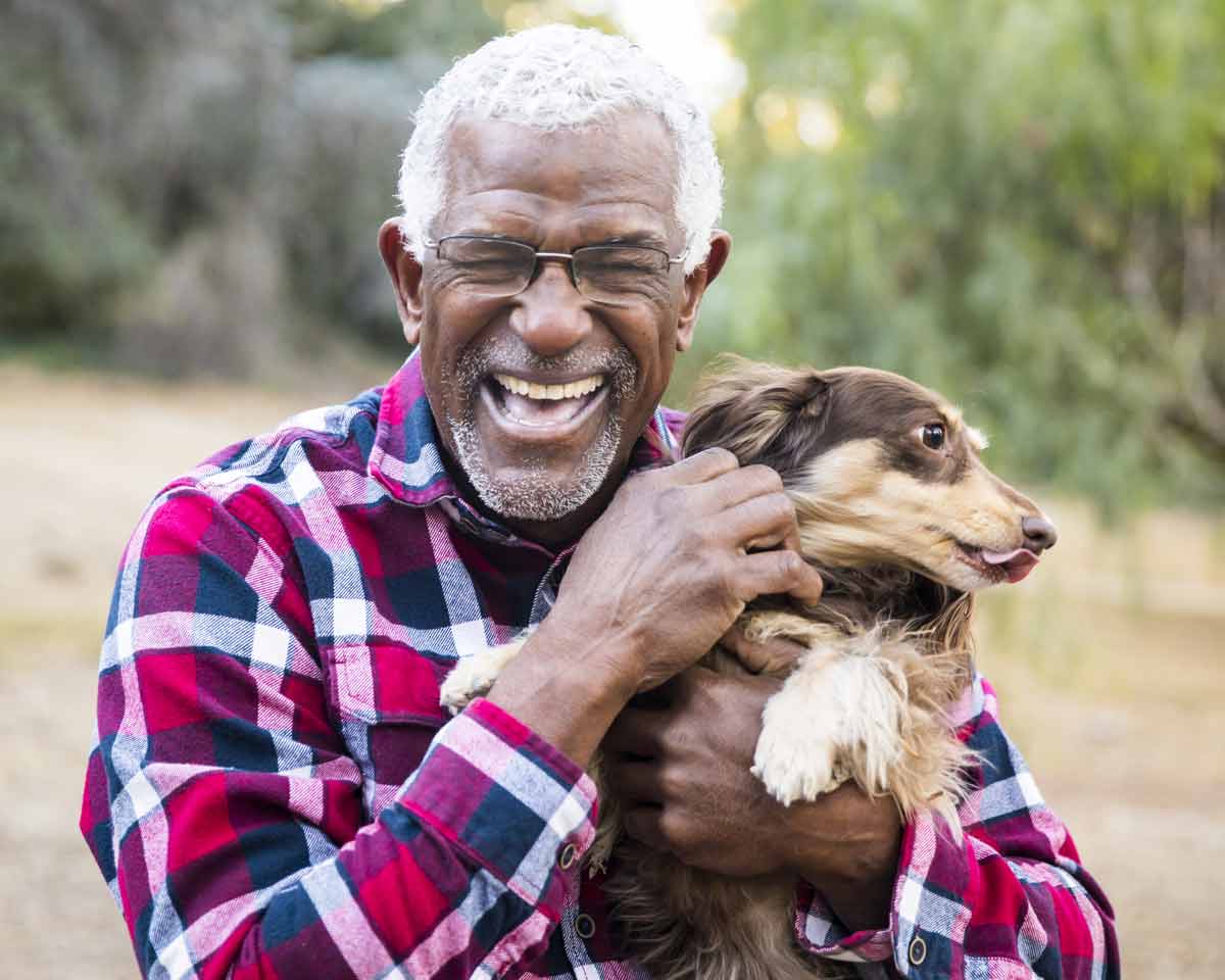 Male assisted living resident in flannel shirt and glasses smiles outside and holds dog