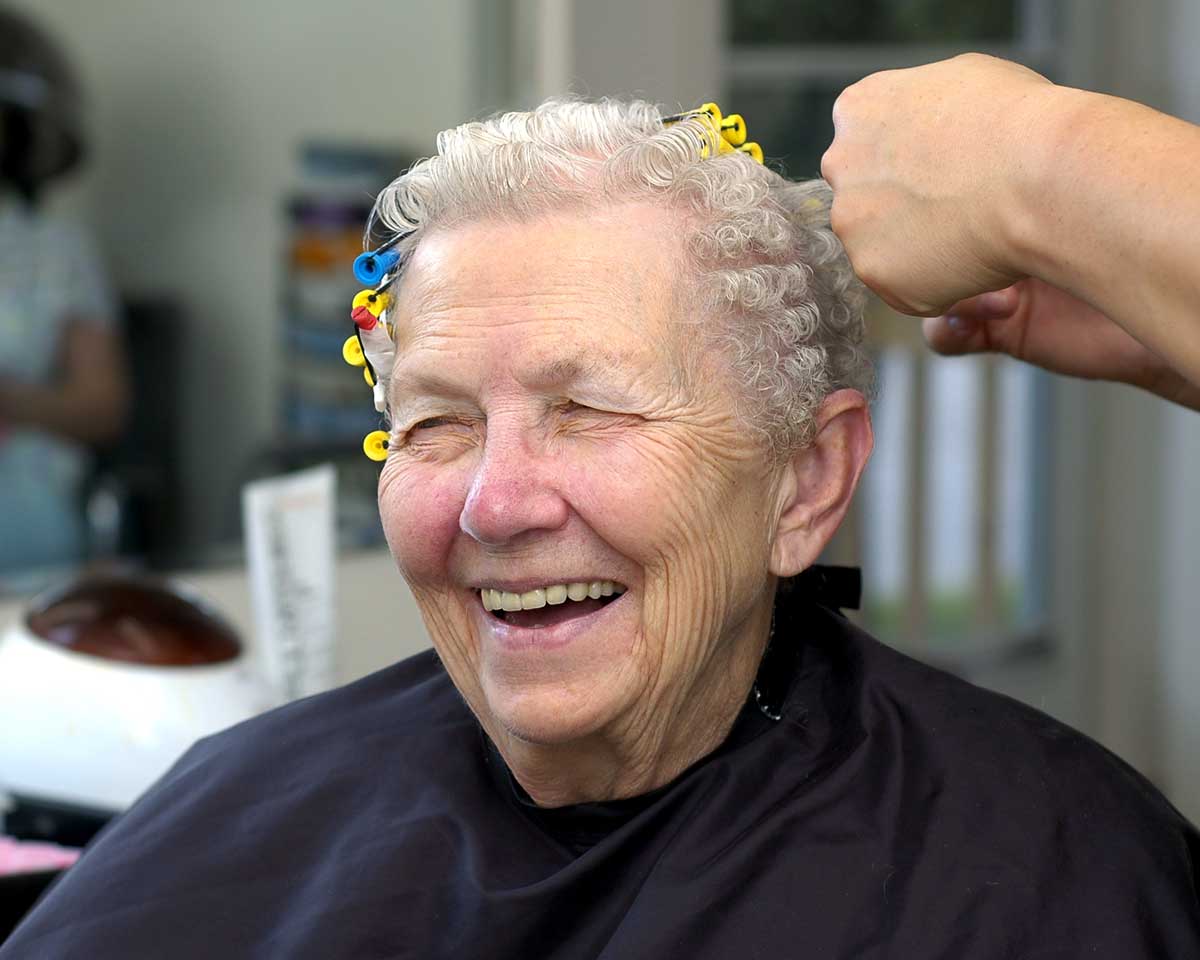 Older female sits in salon chair and wears black cape while having hair styled with curlers
